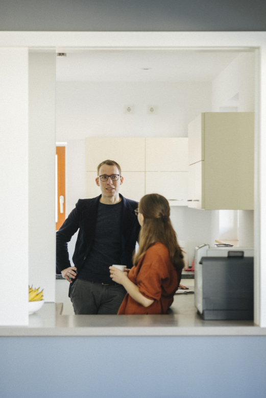 Eine Frau mit einer Tasse in ihren Händen steht vor einer Kaffeemaschine in der Küche im 2nd floor und redet mit einem Mann, der neben ihr steht.