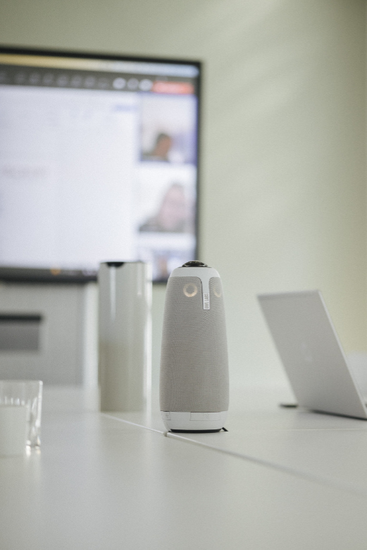 Eine Nahaufnahme der Meeting Owl, die auf einem Tisch steht, daneben ein Laptop. Im Hintergrund läuft eine Videokonferenz auf einem Bildschirm.